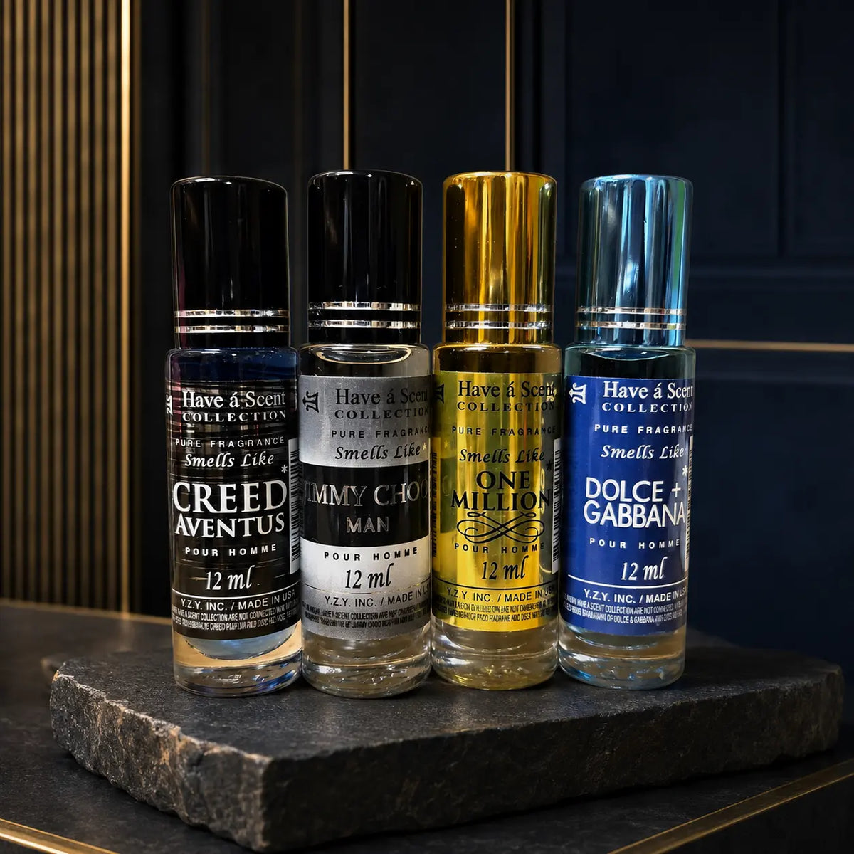 Four cologne bottles from Have & Scent Collection lined up on a dark stone slab. The bottles feature black, silver, gold, and blue metallic caps and labels, representing Creed Aventus, Emmy Chou Man, One Million, and Dolce & Gabbana.