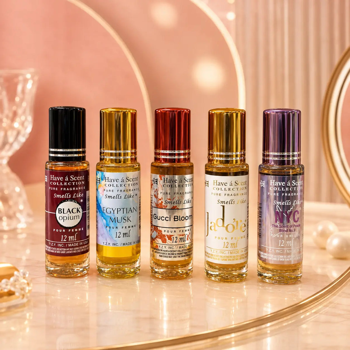 Five small perfume rollerball bottles from Have & Scents Collection are lined up on a reflective marble surface with a warm pink circular backdrop. The bottles feature black, gold, bronze, and purple caps and display scents including Black Opium, Egyptian Musk, Gucci Bloom, La Dolce Vita, and New York NY.
